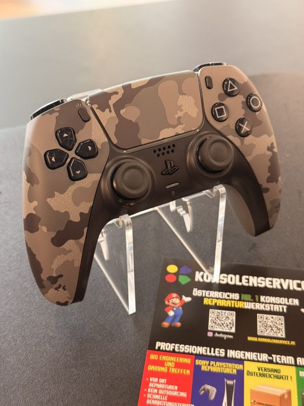 SONY DualSense® Wireless-Controller – Gray camouflage • Konsolenservice.at Close-up of a PlayStation controller with desert camouflage skin on a display stand, showing left thumbsticks and four action buttons on the right.